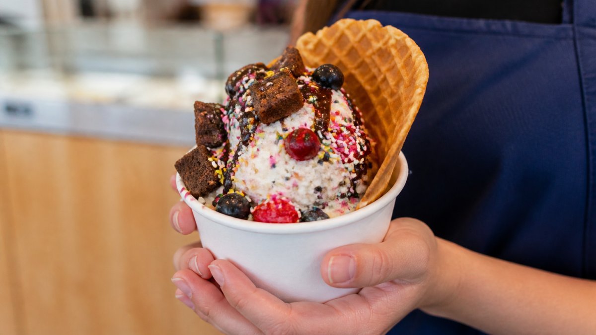 Close-up of hands holding a custom frozen custard cup topped with colorful mix-ins, fresh toppings, and a homemade cone, showing creamy texture and vibrant presentation under warm lighting